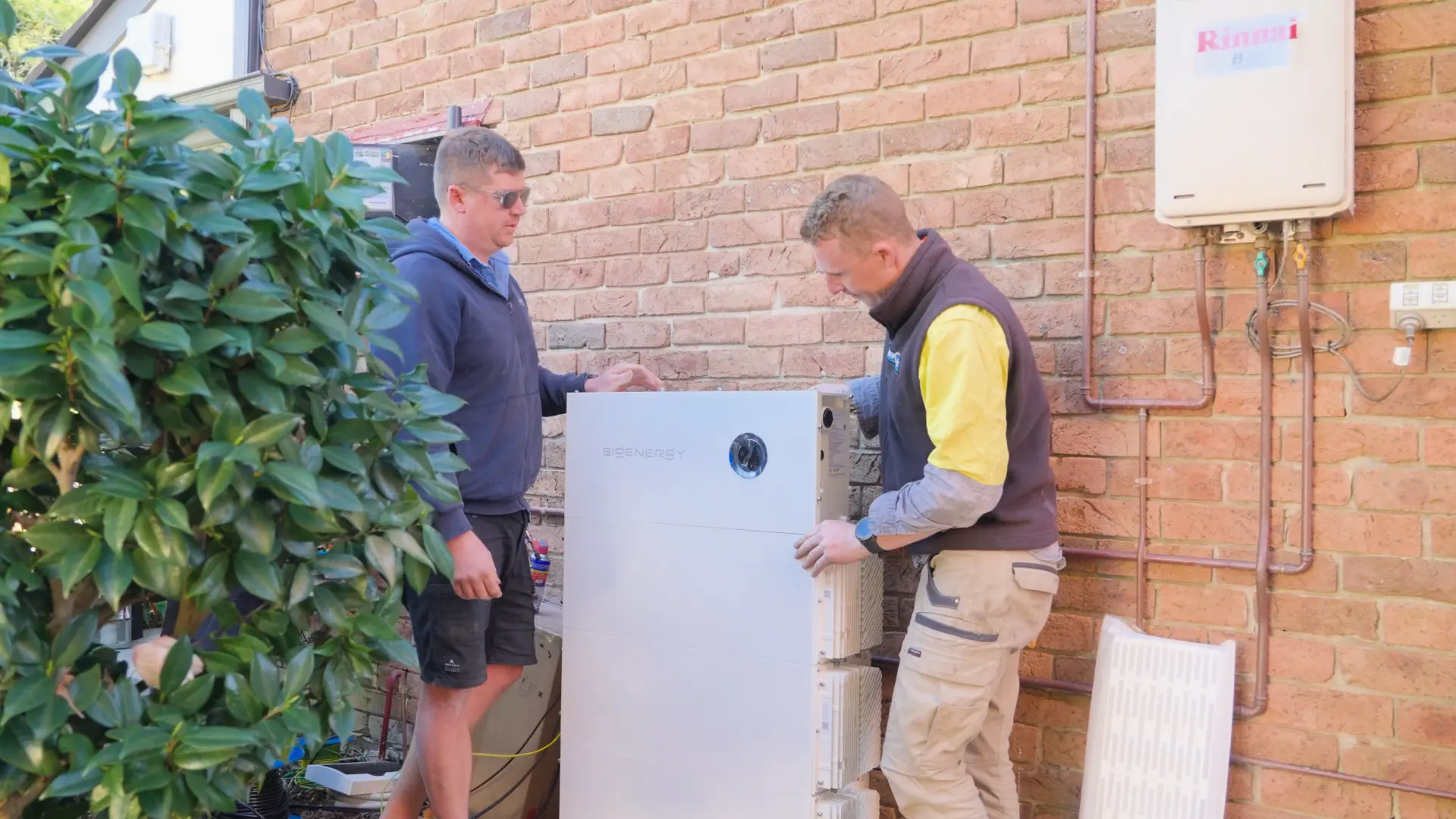 Two SCE installers positioning a Sigenergy battery against a brick wall